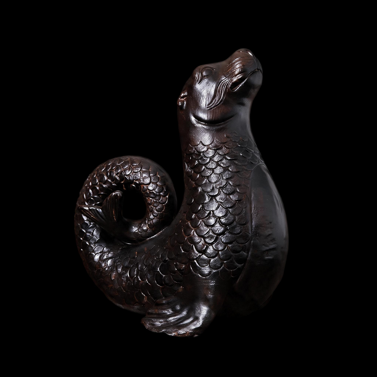 Surreal black sculpture of a seal with a scaled body and coiled fish tail, head lifted